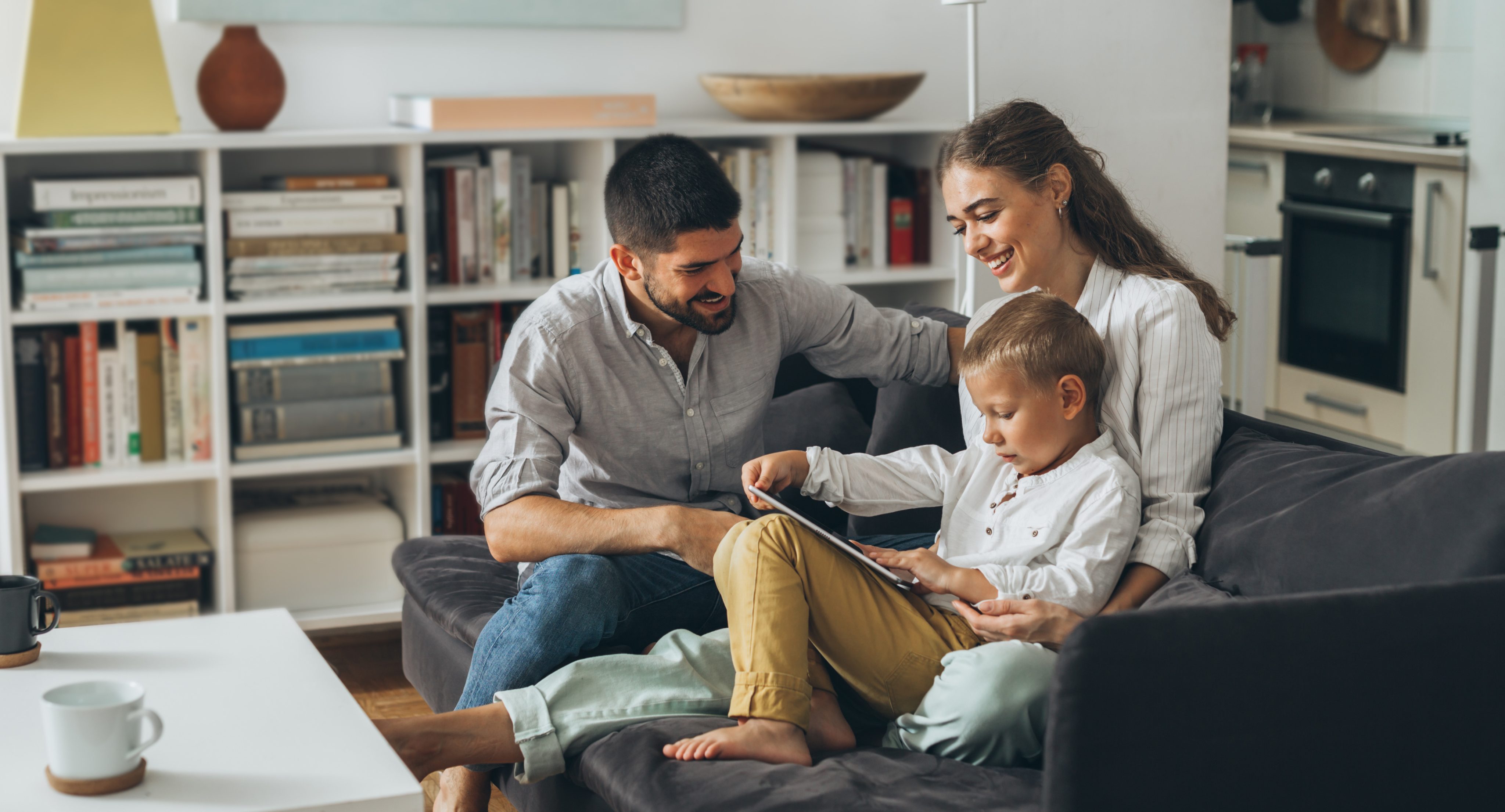 Family of three sitting on a gray couch smiling and looking at the boy's tablet with bookshelves behind