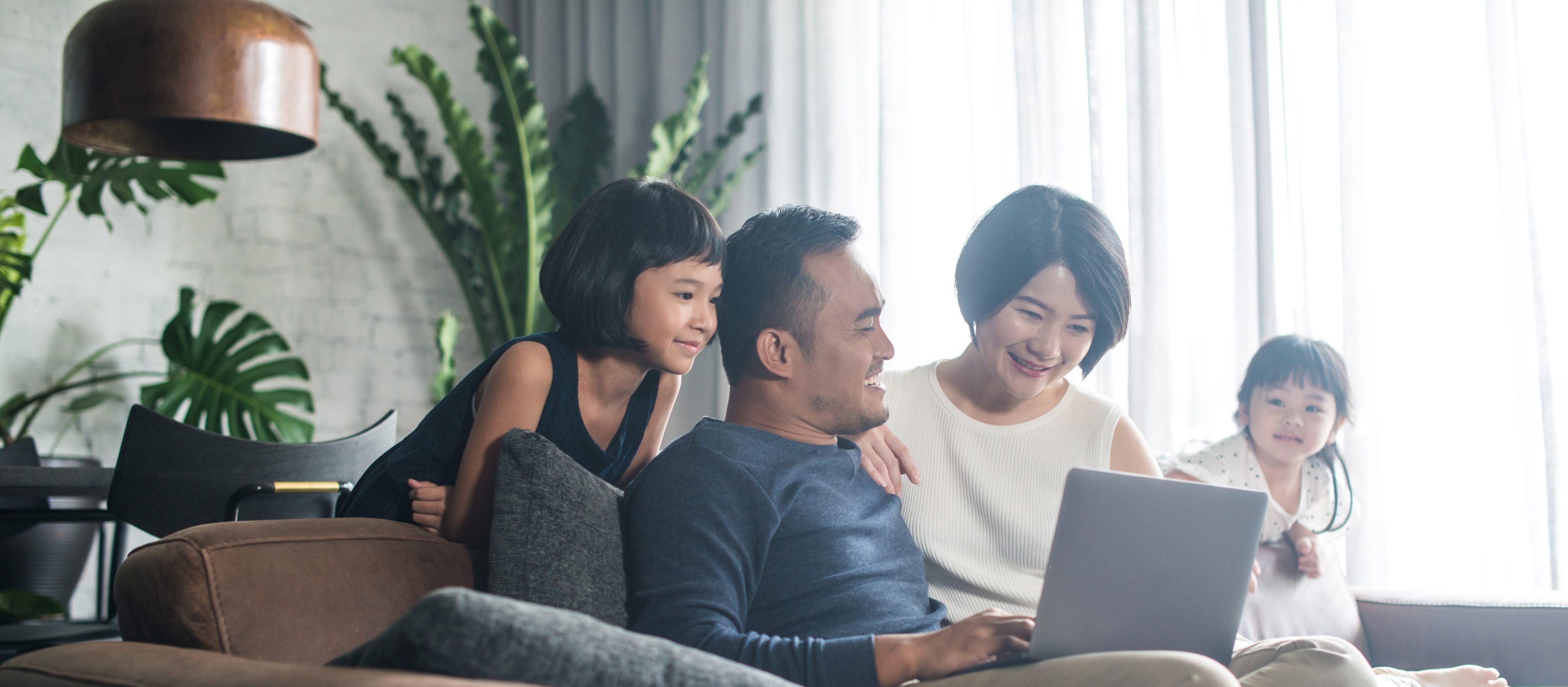 Family of four sitting on a brown couch looking at a laptop in the dad's lap with a large window and white brick wall behind